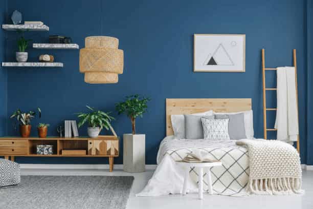 A modern bedroom with a bold navy blue accent wall, featuring a wooden bed frame with grey and white patterned bedding. The room is decorated with a woven pendant light, floating marble-style shelves, and various indoor plants on a wooden console.