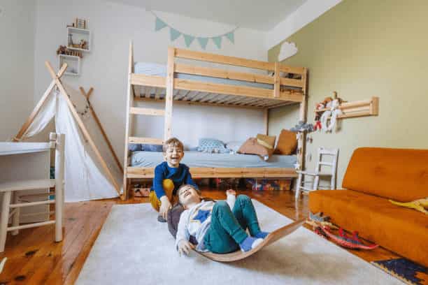 Two young children playing on a wooden balance board in a bright bedroom featuring a wooden bunk bed, a small white teepee, and a sage green accent wall.