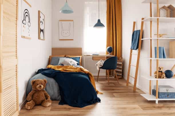 A cozy, narrow kids' bedroom with a navy blue and mustard yellow bedspread, a teddy bear on the floor, and white shelving units against wood-look flooring.