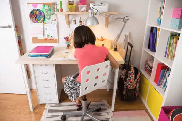 A young girl sitting at a white desk in a well-organized study area with colorful wall decorations, a white bookshelf, and bright storage bins.