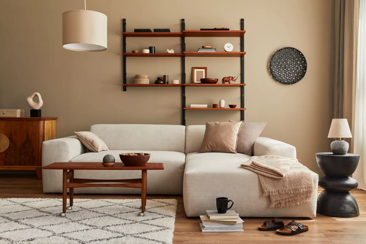 A modern light brown living room featuring a cream sectional sofa, minimalist wooden shelving with decor, and a mid-century coffee table on a geometric rug.