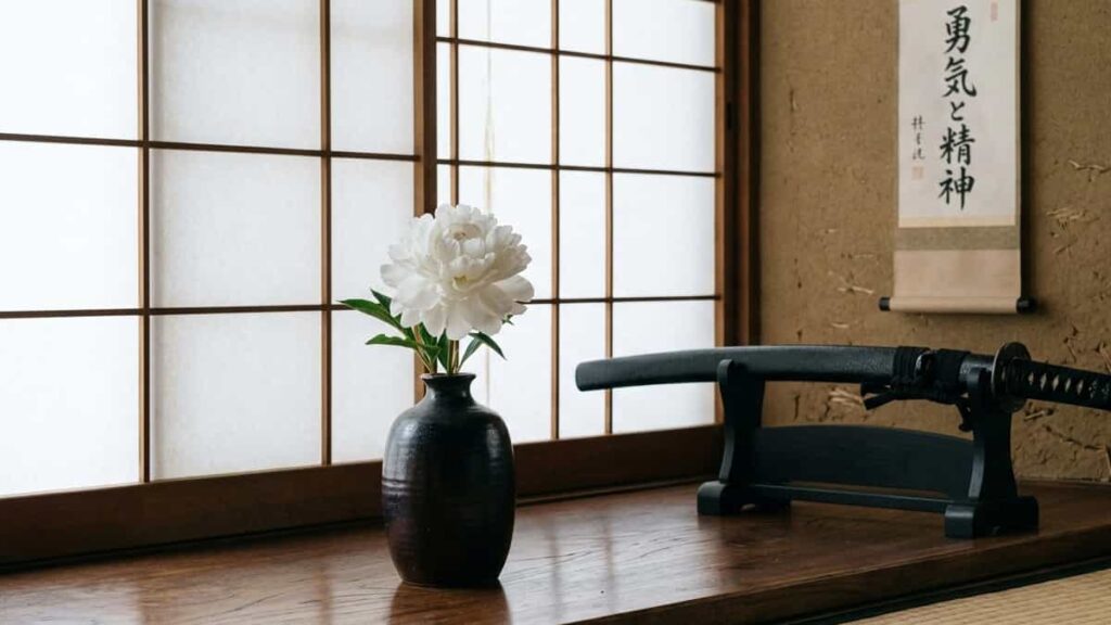 Single white peony in a lacquered Japanese vase beside a samurai katana and calligraphy scroll.