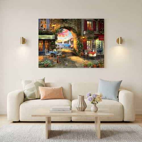 Cozy living room with an off-white sofa and pastel designer cushions featuring a large Mediterranean harbor sunset canvas print. Two gold wall sconces illuminate the artwork on an off-white wall above a light wood coffee table.
