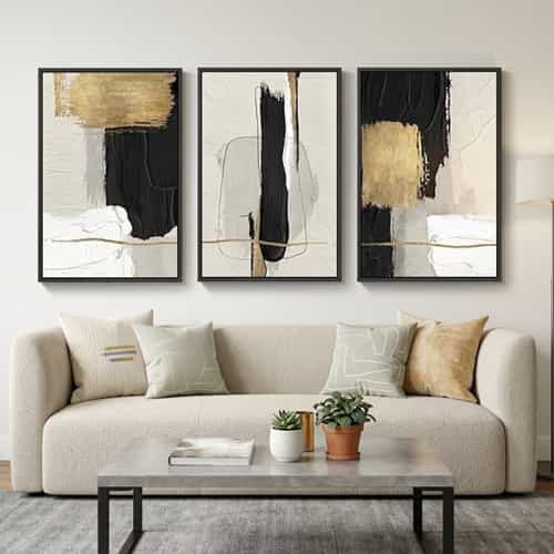 Minimalist living room featuring a cream NY style rounded sofa with light-colored abstract pillows, a marble coffee table with two small potted plants, and a three-piece black and gold abstract wall painting on an off-white wall.