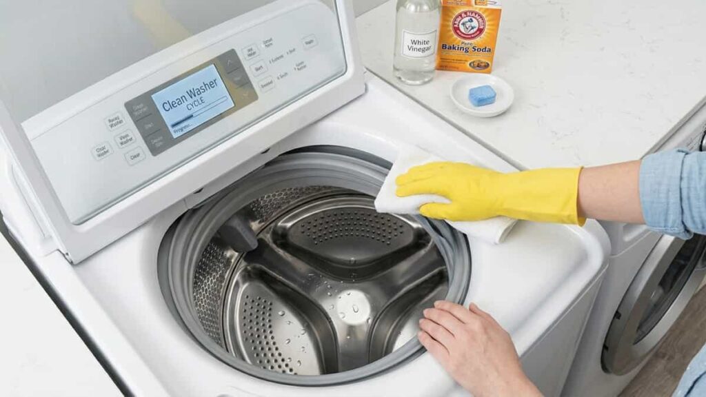 Person in yellow glove wiping the rubber gasket of a clean front load washing machine drum with vinegar and baking soda supplies on top.
