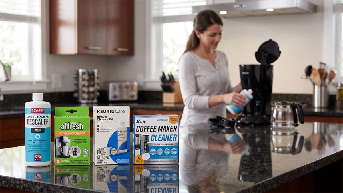 Four coffee maker descalers (IMPRESA, Affresh, Keurig, and ACTIVE) lined up on a dark granite countertop with a woman cleaning a coffee machine in the background.